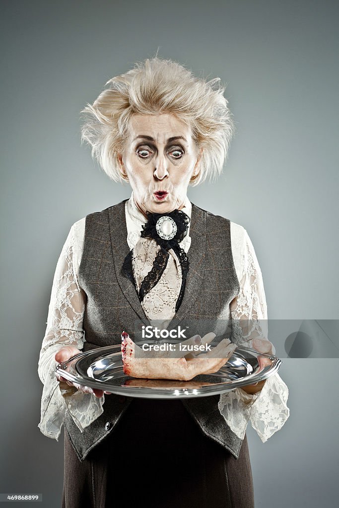 Spooky senior lady Contemporary portrait of victorian style spooky countess serving a human hand on the tray. Studio shot. Bizarre Stock Photo