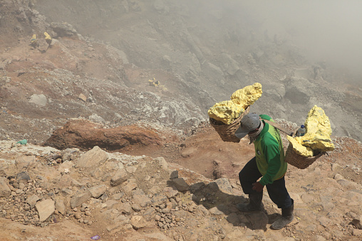 Kawah Ijen, Indonesia - August 8, 2011: Miner carries baskets with sulphur in the fumes of toxic volcanic gas from the sulphur mines in the crater of the active volcano of Kawah Ijen, East Java, Indonesia.
