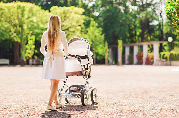 family, child and parenthood concept - happy mother walking with baby stroller in park from back