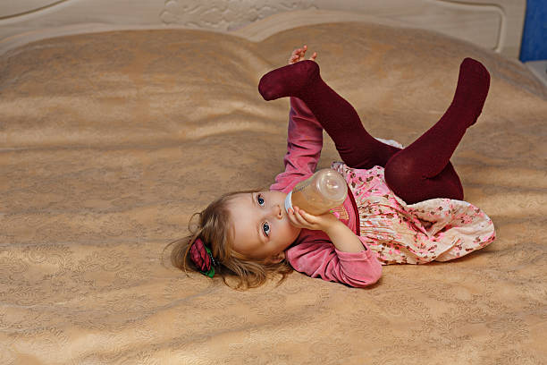 Little cute girl drinking from a bottle lying on the bed in the bedroom.