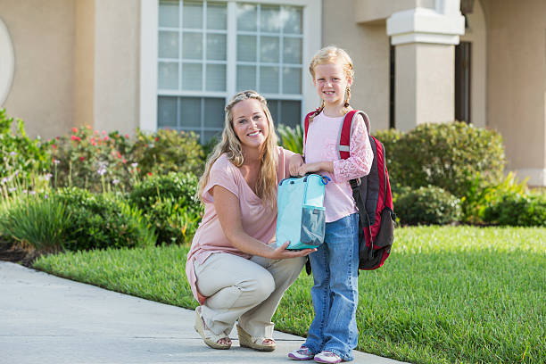 A little girl standing with her mother in front of their house, ready for school, carrying a lunchbox and backpack.  They are smiling at the camera.