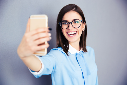 Laughing businesswoman making selfie photo on smartphone. Wearing in blue shirt and glasses. Standing over gray background