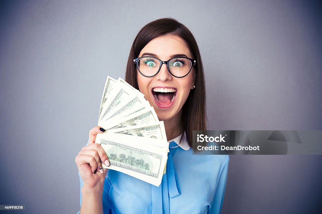 Businesswoman holding bill of dollars and shouting Laughing businesswoman holding bills of dollar and shouting over gray background. Wearing in blue shirt and glasses. Looking at camera Currency Stock Photo