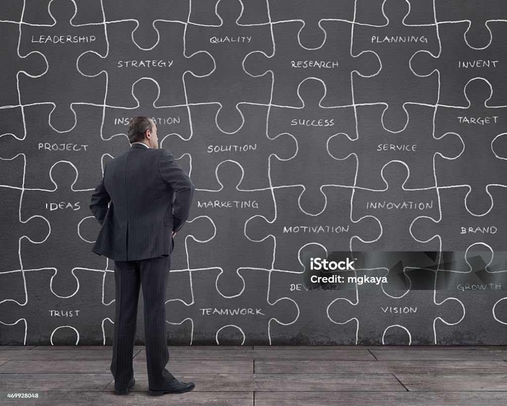 Businessman with Jigsaw Puzzle and Business Terms Rear view of businessman standing on wooden floor with jigsaw puzzle sketched (chalk drawing) and business terms on the wall. He is looking further, thinking, searching new ideas. Exploration Stock Photo