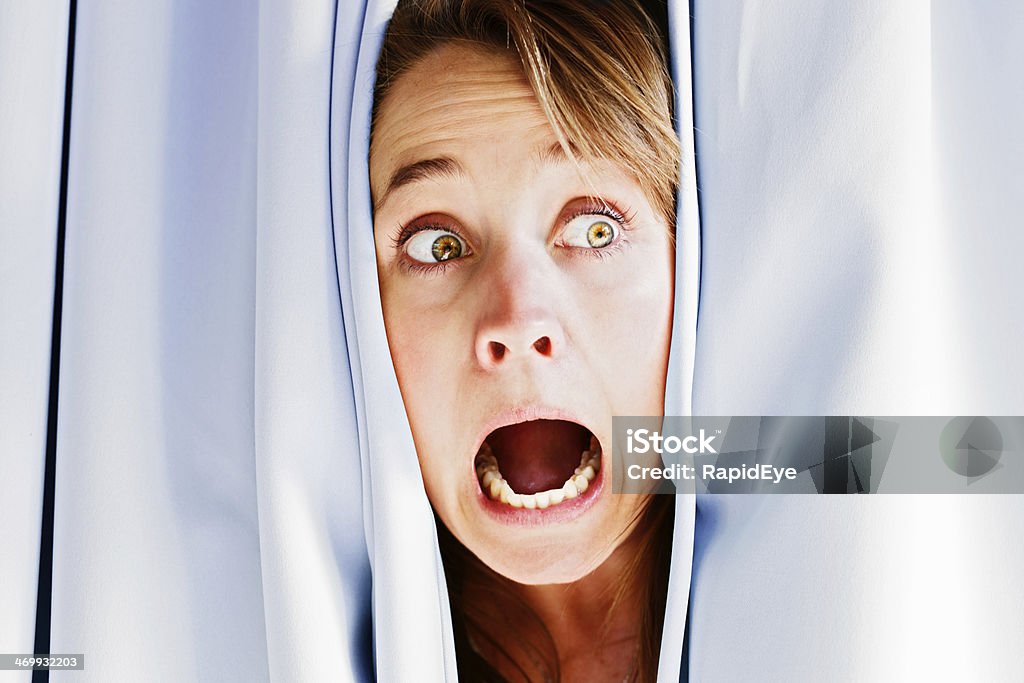 Beautiful blonde peeping through curtains screams in terror Peeping sideways through closed drapes, this pretty blonde screams in terror. Could be burglars or she Curtain Stock Photo