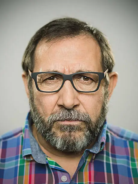 Portrait of a real spanish man with negative expression. Studio photography with sharp focus on eyes. The man wears modern glasses and shirt and has a grey beard.