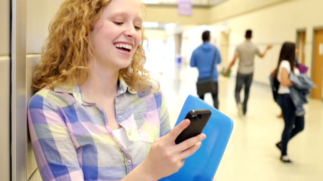 Teen girl laughing as she texts on smart phone in high school hallway