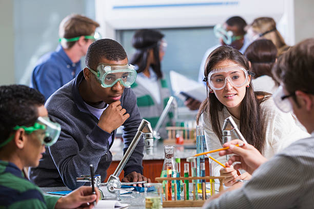 Young adult students of various ethnic backgrounds doing a chemistry experiment in class. The students are all wearing protective safety goggles.