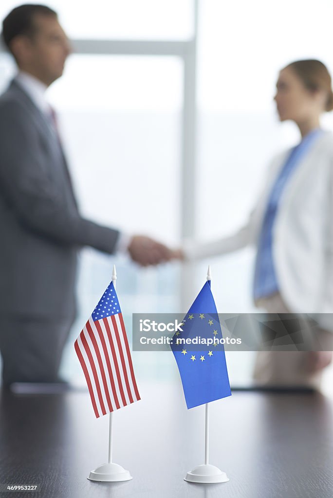 Global partnership American and European Union flags on the table and two people shaking hands in the background Adult Stock Photo
