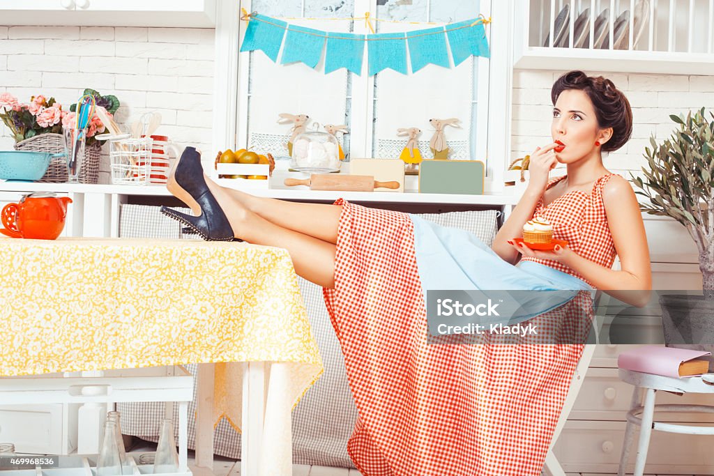 A girl licking her finger in the kitchen Pin-up girl style. Girl sitting on a chair at the table, in the hands holding a muffin. Cake Stock Photo