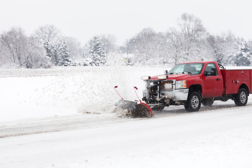 Smaller Truck with a smaller snow plow attached is plowing a country road. Trees line the distance just beyond a field. Snow is falling and the plow is kicking up snow along side the road. Red truck stands out against the white and gray surroundings. Room left for cropping or copy. XXXL size.