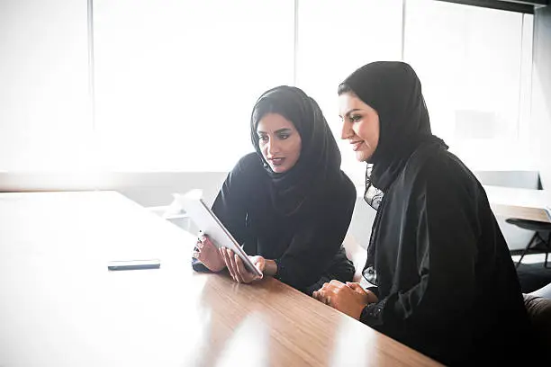 Candid portrait of two arab women using digital tablet at open plan desk in modern workplace. They are wearing traditional black abaya in contemporary business office with window behind. Dubai, United Arab Emirates, UAE.