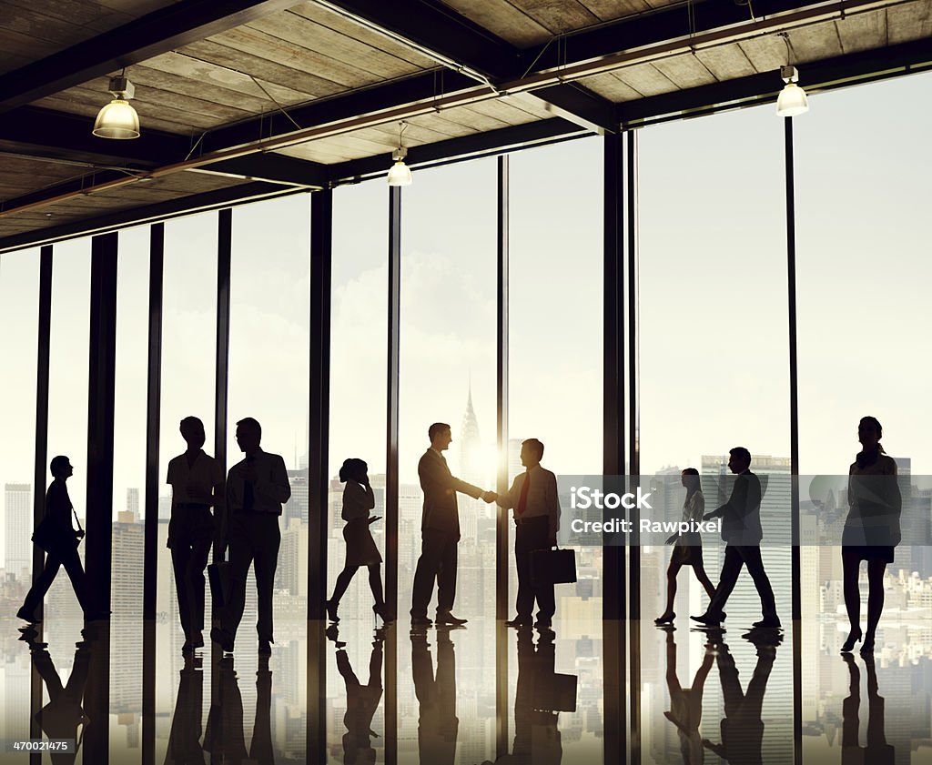 Silhouette businesspeople entering office building Silhouettes of business people walking, talking, and shaking hands with glass windows and a view of a cityscape in the background. Business Person Stock Photo