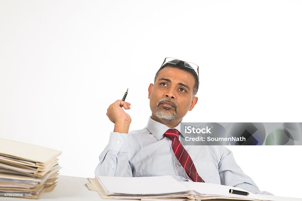Mature businessman lost in deep thoughts at his desk. Mature businessman working at his desk and thinking. He is holding a pen in his hand and looking upwards. He has his laptop in front of him. He is deeply engrossed in his work and has a serious face. He is in business attire and wearing a red neck-tie, Contemplation Stock Photo