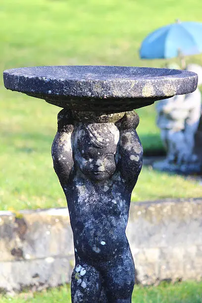 Photo showing an old moulded stone birdbath, where the dish / saucer of water is supported by a small boy statue.  The cherub-like figure is shown supporting the dish, which is being balanced on his head.