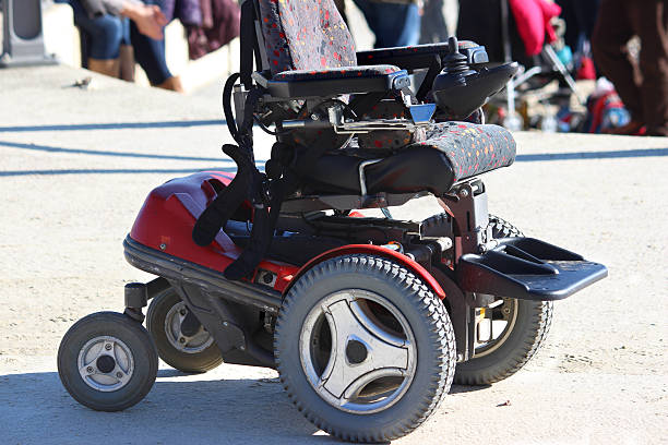 Photo showing a modern electric wheelchair, which has two large wheels at the front and two smaller wheels at the back.  The motor sits to the rear of this red powered wheelchair, being operated by controls next to the arms of the seat.  A joystick provides the disabled passenger with an easy way to control the steering mechanism.