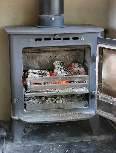 Photo showing a small cast iron woodburner, which has been pictured with its door open to reveal the logs, ash and warming fire burning inside.  Metal wood burning stoves are often referred to as multi-fuel stoves, since they are able to burn either wood or alternatively, a smokeless fuel, such as coke, coal or charcoal options.