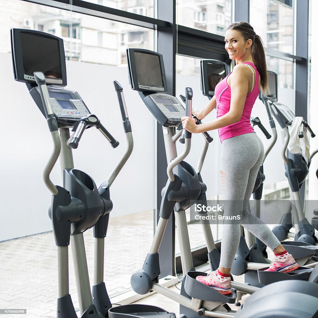Smiling woman exercising on cross trainer in a health club. Happy woman doing leg exercises on cross trainer in a gym and looking at the camera. One Woman Only Stock Photo