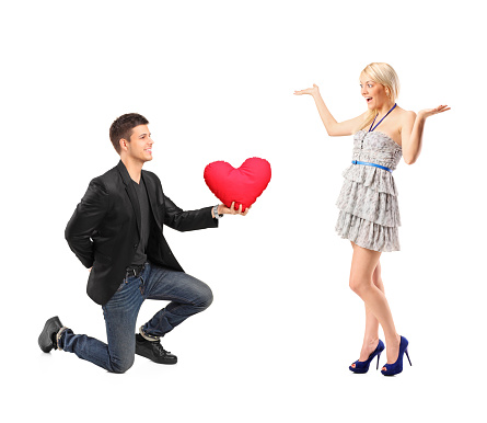 A romantic man on his knees holding a red heart shaped pillow and an excited blond woman isolated on white background
