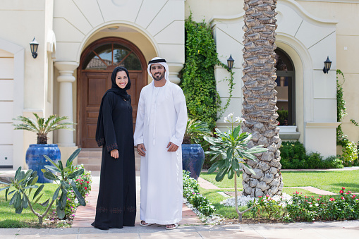 Middle Eastern couple standing in front of their house on a sunny day, both wearing the traditional Emirati dresses .