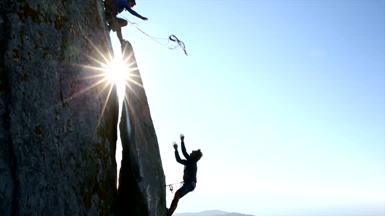 Climber throws rope to teammate, on cliff above hills, sea