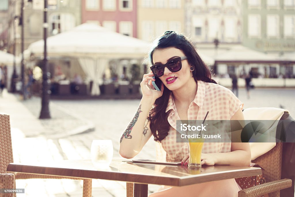 Young woman talking on phone outdoor Young hipster woman with tatoo on her arm wearing sunglasses sitting at the table in outdoor restaurant in the city and talking on phone. Orange juice on the table. Summer time.  2015 Stock Photo