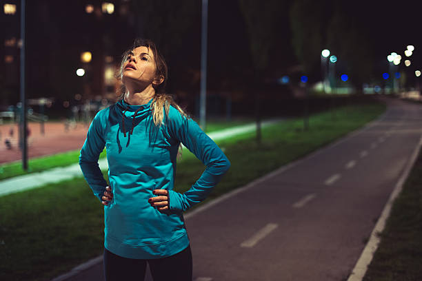 After running Young tired woman standing on the running track outdoor after running. It's night time. tired after work stock pictures, royalty-free photos & images