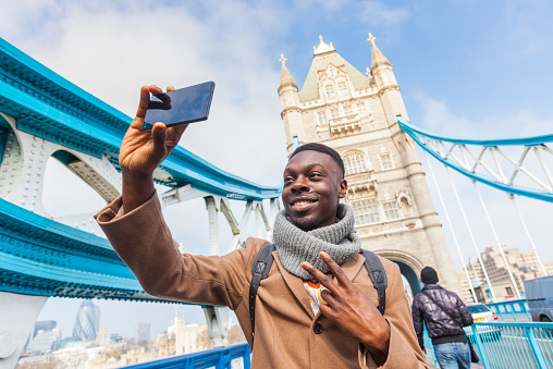 Smiling black man taking selfie in London with Tower Bridge on background. He is holding the phone and looking at camera. Photo taken on a sunny winter day.