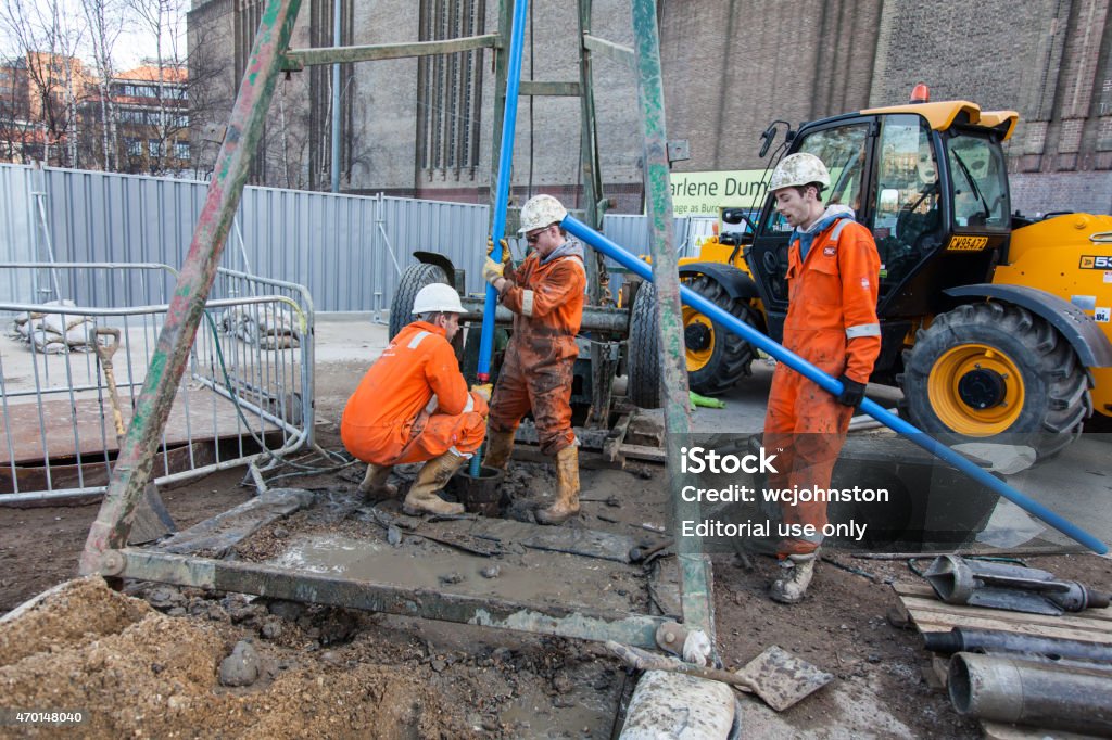 Workmen in orange overalls London, England - March 10, 2015: Workmen in orange overalls and white hardhat outside the Tate Gallery in London holding a blue water pipe. one has yellow gloves and the other black. they are next to a borehole drilling rig. and behind is a JCB. a fence can be seen. One man is croched down the other stands up. another stands to the right holding another pipe. 2015 Stock Photo