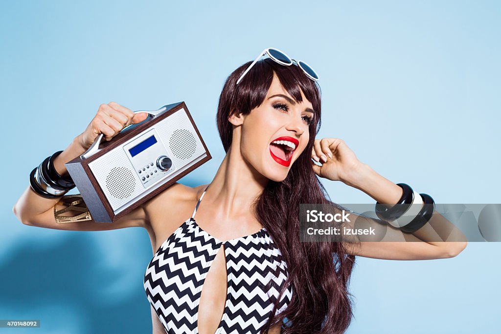 Excited young woman wearing swimsuit, listening to the radio Summer portrait of glamour brown long hair young woman wearing black and white swimsuit holding small radio on her shoulder, listening to the music and singing. Standing against blue background. Studio shot, one person. Radio Stock Photo