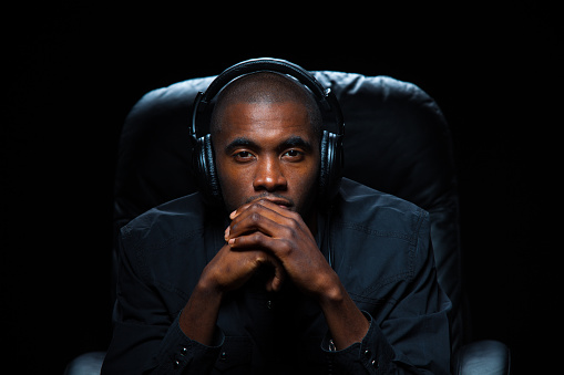 A young man gazes intently at the camera, hands clasped at his chin, with over-ear headphones on. He is leaning forward in a black leather chair in a darkened room, in an atmosphere of intense concentration.