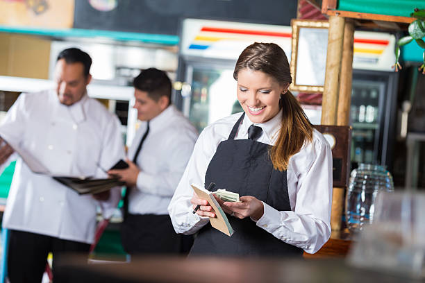 Friendly waitress in Tex-Mex restaurant counting tips after shift Pretty mid adult Caucasian woman is standing in front of restaurant commercial kitchen. She is wearing a white shirt with a black tie and apron, and is a professional waitress. She's smiling while looking down and counting cash tips after working long shift. Chef and restaurant manager are standing in background of casual dining restaurant. mex mexican restaurant tex stock pictures, royalty-free photos & images