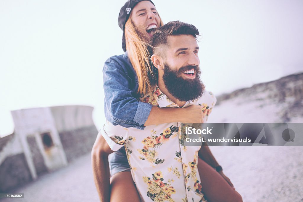 Girl laughing while hipster boyfriend piggybacks her at the beac Happy girl laughing loudly while her hipster boyfriend is piggybacking her at the beach Couple - Relationship Stock Photo
