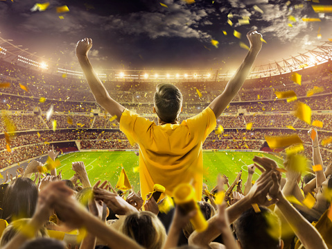 Crowd of sports fans cheering during a match in stadium. Excited people standing with their arms raised, clapping, and yelling to encourage their team to win.