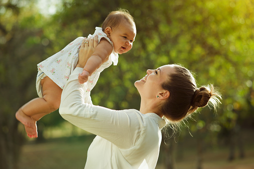 Beautiful woman lifts high her adorable baby up mid air and looks at her smiling. Happy parent spending time playing with daughter in park at sunset. Medium shot