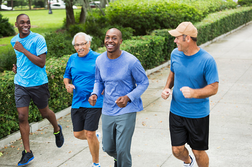 Multi-ethnic group of people running or power walking in a park.  The four men are wearing blue shirts, running for a common cause, maybe cancer awareness.  Focus is on the young African American man looking at the camera.