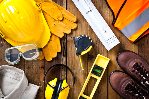 Personal protective workwear and blueprint with some measuring instruments shot directly from above on rustic wood background. The protective workwear includes hard hat, gloves, earmuff, goggles, steel toe shoes, and safety vest. The composition also includes a tape measure and bubble level and a construction blueprint, all items used by construction worker or engineer. Predominant colors: yellow and brown.