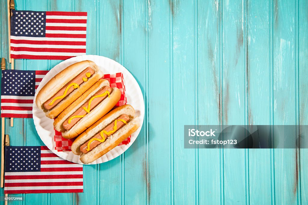 Summer holiday picnic. Hot dogs, USA flags. Blue wooden table. Happy Independence Day America!  Three USA flags form a left side border with hot dogs on white plate. Copyspace to right. Teal blue wooden beadboard background with grunge painted effects.  Memorial Day, July 4th, Veteran's Day, Labor Day, Flag Day summer picnic concepts.  American pride and patriotism. Barbecue - Meal Stock Photo