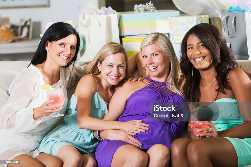 Expectant mother at baby shower with friends A group of multi-ethnic, mid adult women throwing a baby shower for their pregnant friend, sitting on a couch laughing and having fun.  Presents and gift bags are on table behind them.  They are smiling at the camera.  The expectant mother is wearing a purple dress. 2015 Stock Photo