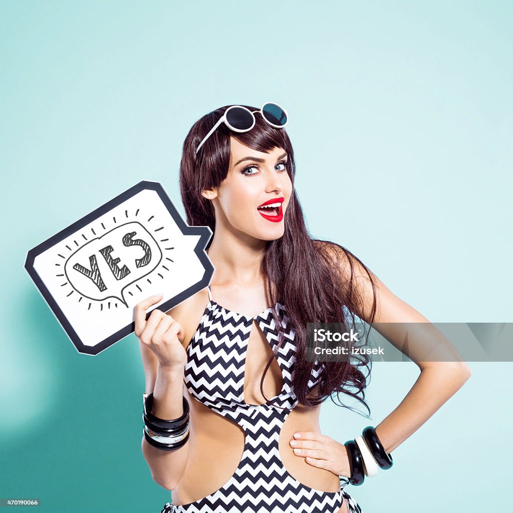 Happy young woman wearing swimsuit holding speech bubble Summer portrait of surprised brown long hair young woman wearing fashionalbe black and white swimsuit. Standing against turquoise background and holding speech bubble with text yes in hand, looking at camera. Studio shot, one person. Blue Background Stock Photo