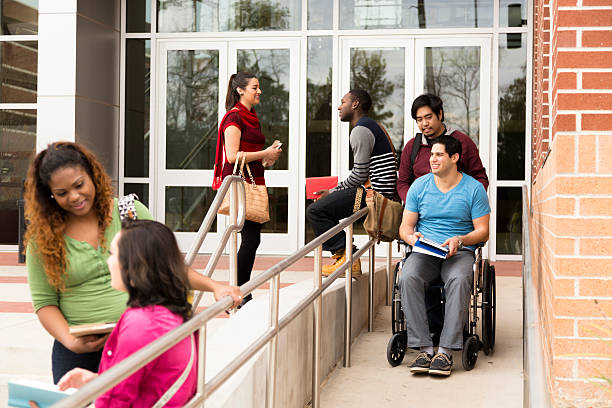 Education: Disabled student helped down wheelchair ramp. College campus. Wheelchair-bound college student is helped down the ramp by a friend or volunteer.  Other students in background talk on campus.  accessibility for persons with disabilities photos stock pictures, royalty-free photos & images