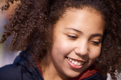 Close-up of a cheerful and happy teenage girl with a toothy smile, looking down. The girl is of mixed race with dark skin and brown, curly hair.