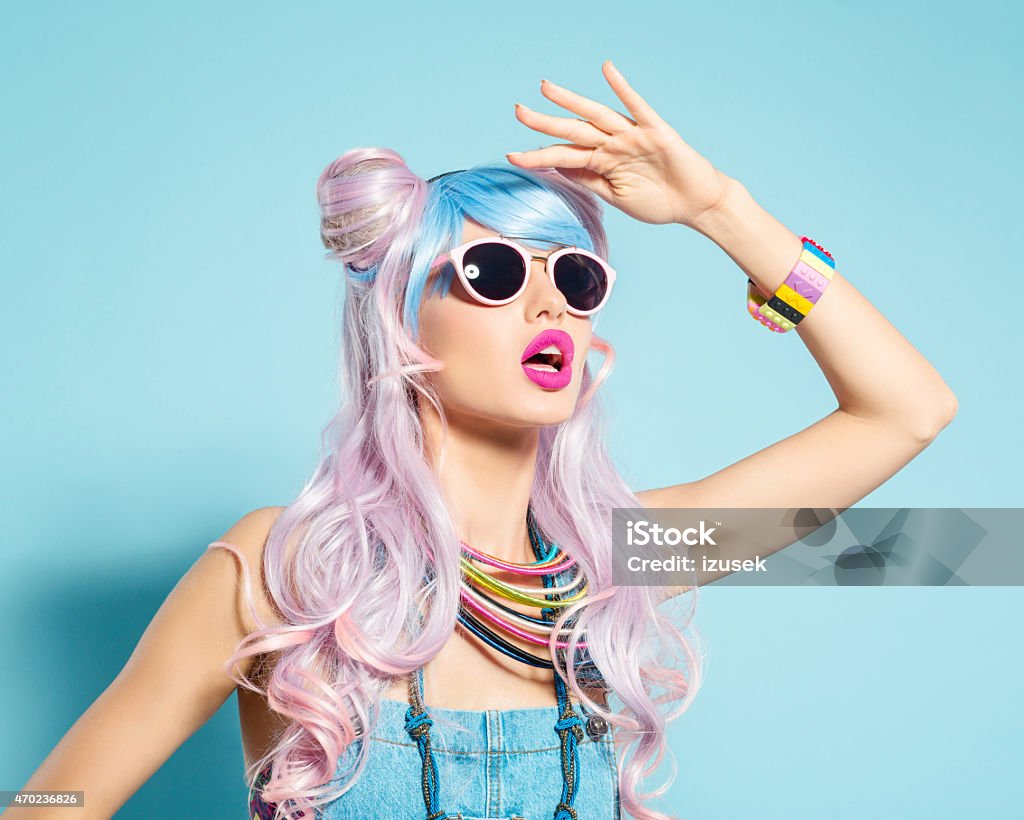 Pink hair girl in funky manga outfit Portrait of sensual manga style blue-pink hair young woman wearing denim coveralls and sunglasses. Standing against blue background, looking away with raised hand. Studio shot, one person. Women Stock Photo