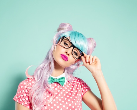 Portrait of confident manga style blue-pink hair young woman wearing pink polka dot dress with collar, bow tie and nerd glasses. Standing against turquoise background, looking at camera. Studio shot, one person.