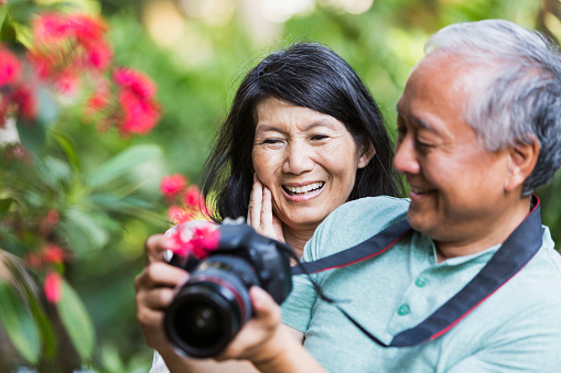 Close up of a senior Japanese couple taking photos with a camera in the park.  The man is holding the camera, but the focus is on his wife who is standing behind him.  They are photographing nature, surrounded by lush foliage and pink flowers.