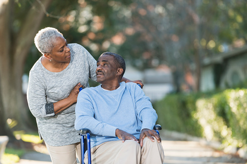 A senior African American couple taking a walk in the park.  The man is sitting in a wheelchair, looking over his shoulder at his wife, who is standing behind him looking at his face.  They are talking, with serious expressions on their faces.  They are wearing casual clothing - tan pants and blue and gray sweatshirts.