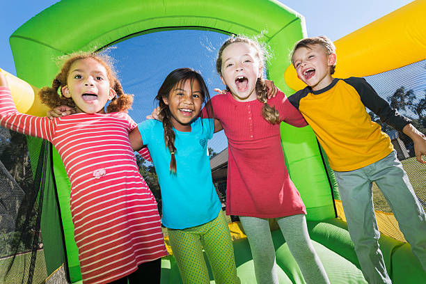 Four multi-ethnic children playing on bouncy castle A group of four multiracial friends having fun on a bounce house.  They are standing side by side with their arms around each other's shoulders, laughing and shouting at the camera.  There is one boy and three girls, 6 to 8 years old.  The inflatable castle is bright yellow and green. fete stock pictures, royalty-free photos & images