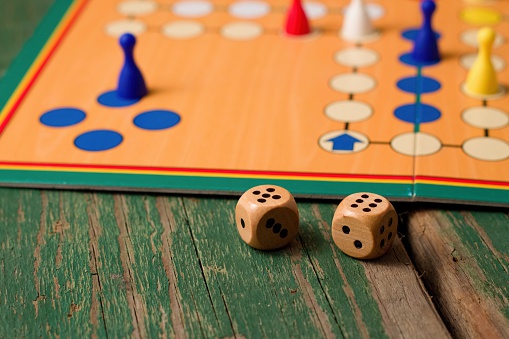 Horizontal photo of Two wooden dices in front of ludo with figurines. Dices have numbers five and six up and all is placed on green old worn wooden board.