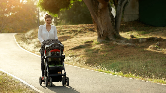 Beautiful woman walking down a lane in park with her little daughter and pushing pushchair, talking to her baby and smiling. Full length view at sunset