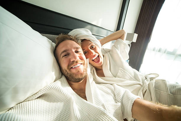 Young cheerful couple taking selfie in hotel room Young funny couple on a comfortable bed in an hotel room taking a selfie in the morning. They both wearing a bathrobe and the woman a towel on her hair. bathrobe photos stock pictures, royalty-free photos & images
